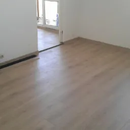 Jasne panele podłogowe ułożone w nowym pomieszczeniu, widoczne paczki paneli przy ścianie, przygotowane do montażu listwy przypodłogowe.