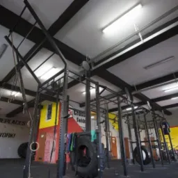 Wnętrze siłowni typu crossfit z czarnymi konstrukcjami do ćwiczeń, oponami i napisem 'Nothing replaces hard work!' na ścianie.