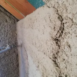 Aplikacja szarego tynku maszynowego na ścianie wewnętrznej, widoczny pistolet natryskowy i fragment drewnianego stropu z niebieskim akcentem.