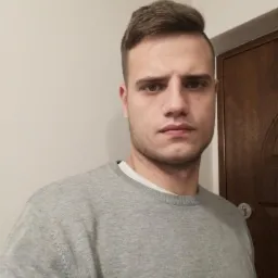 Młody mężczyzna w szarym swetrze, brązowe drzwi w tle, oświetlenie górne. Portret na neutralnym tle.