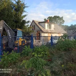 Ciężka maszyna Volvo BL71 na tle częściowo zburzonego domu z odsłoniętą konstrukcją dachu, widoczna przez niebieskie ogrodzenie z siatki.