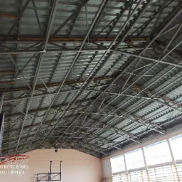 Metalowa konstrukcja dachu hali sportowej z widocznymi elementami podtrzymującymi i oświetleniem, kosz do koszykówki zamontowany na ścianie.