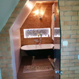 Wykończona łazienka na poddaszu z ceglanymi ścianami, wolnostojącą wanną na złotych nóżkach i dekoracyjną wnęką z podświetlanymi płytkami.