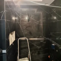 Łazienka w trakcie prac wykończeniowych: czarne płytki imitujące marmur na ścianach, widoczna drabina malarska i krzyżyki dystansowe do płytek, surowa żarówka.