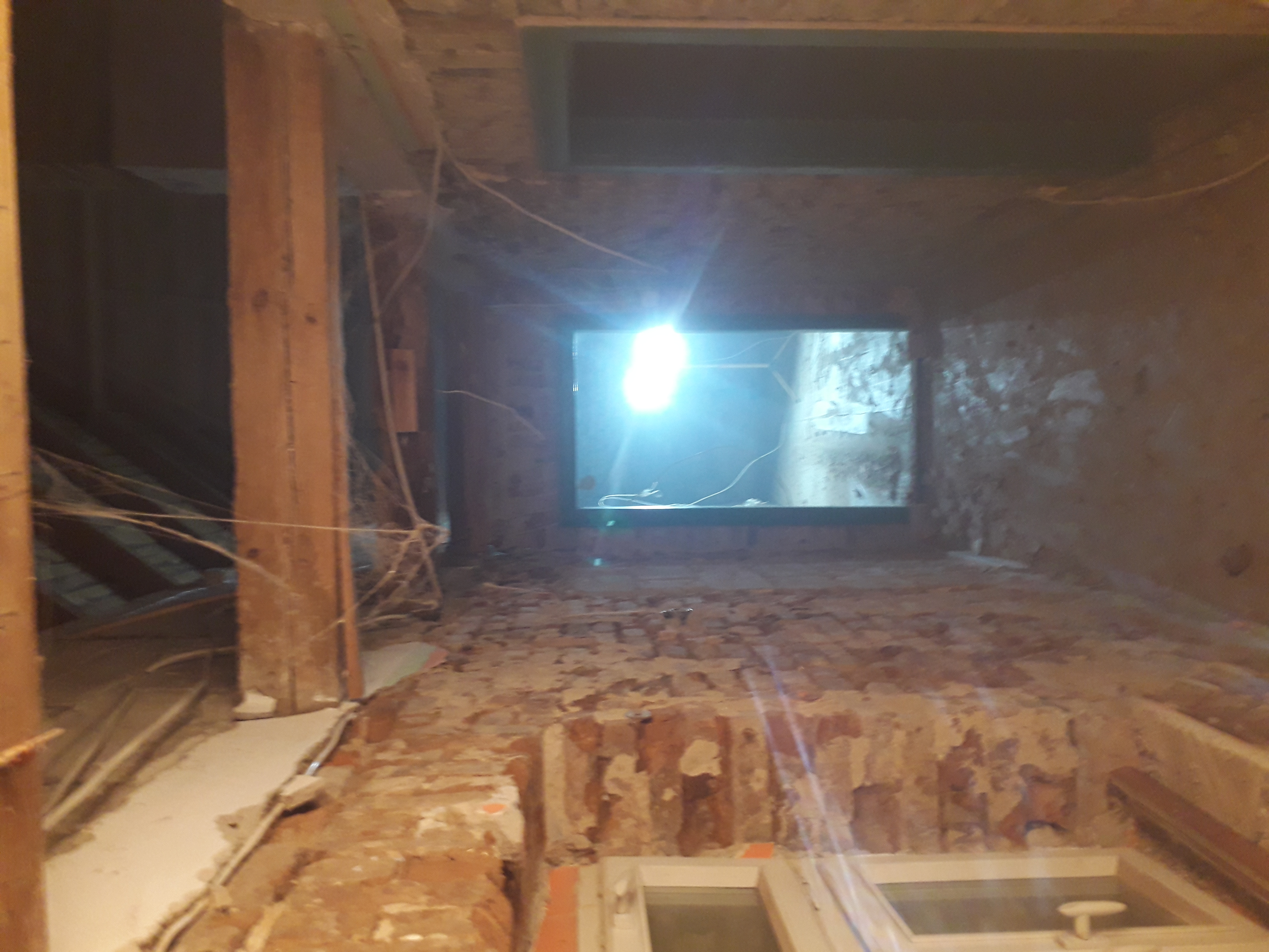 Ceglana ściana w trakcie demontażu z widocznym oknem, drewnianymi belkami i fragmentami instalacji elektrycznej.