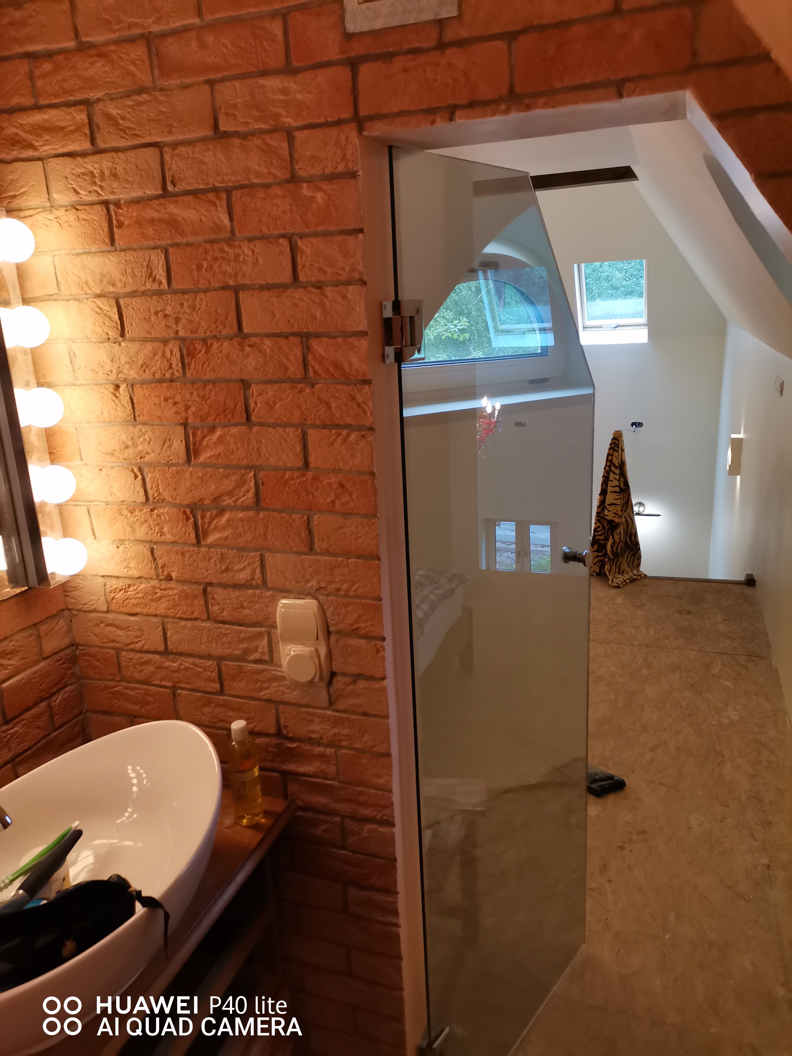 Łazienka w trakcie remontu: umywalka z baterią na tle ściany z cegły, częściowo zamontowana szklana kabina prysznicowa, w tle widoczny strych z oknami dachowymi i podłogą z płyty OSB.