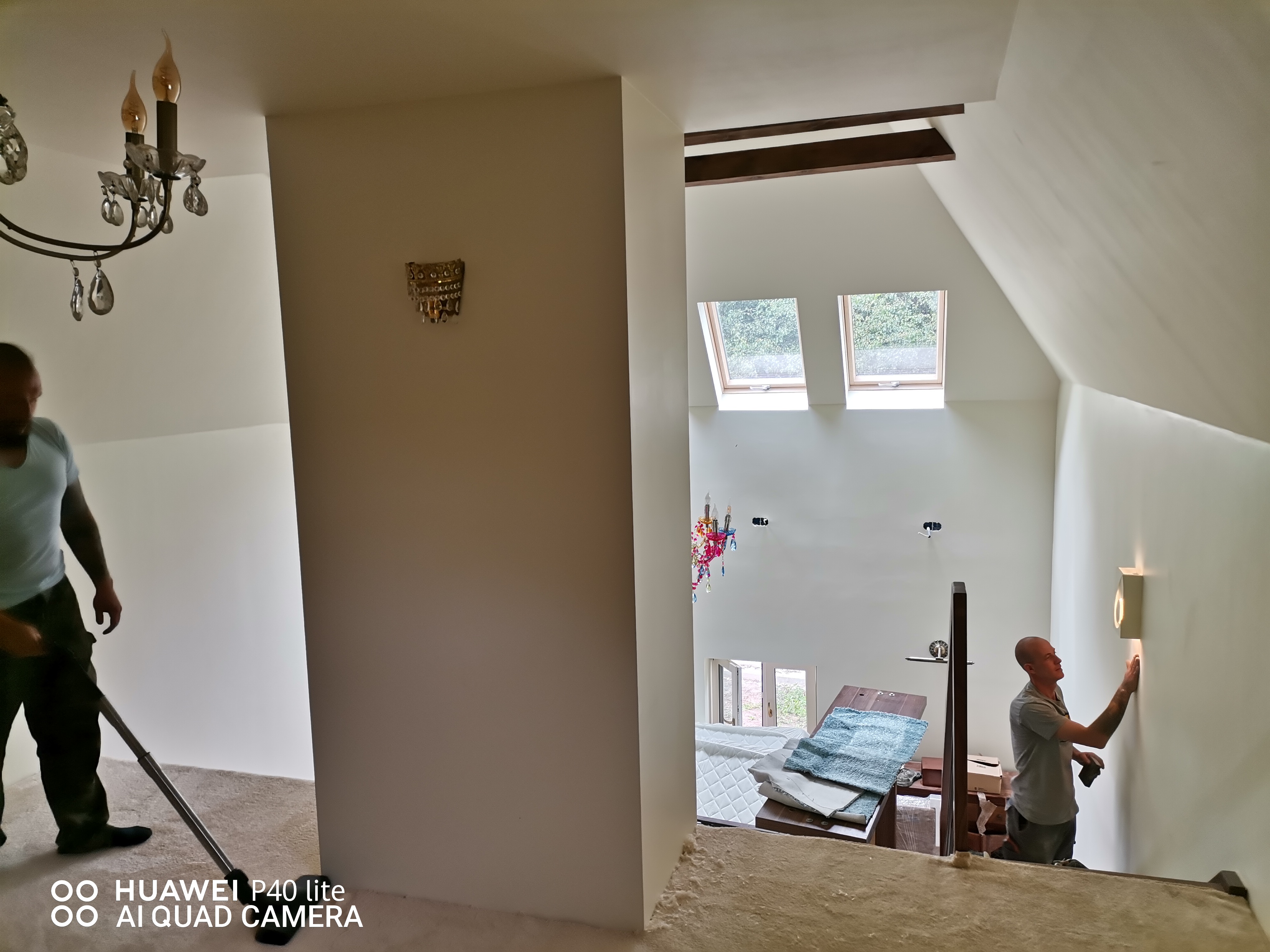 Wnętrze domu w trakcie prac wykończeniowych: pracownik szpachluje ścianę przy schodach, widoczne elementy dekoracyjne, okna dachowe i przygotowane materiały.