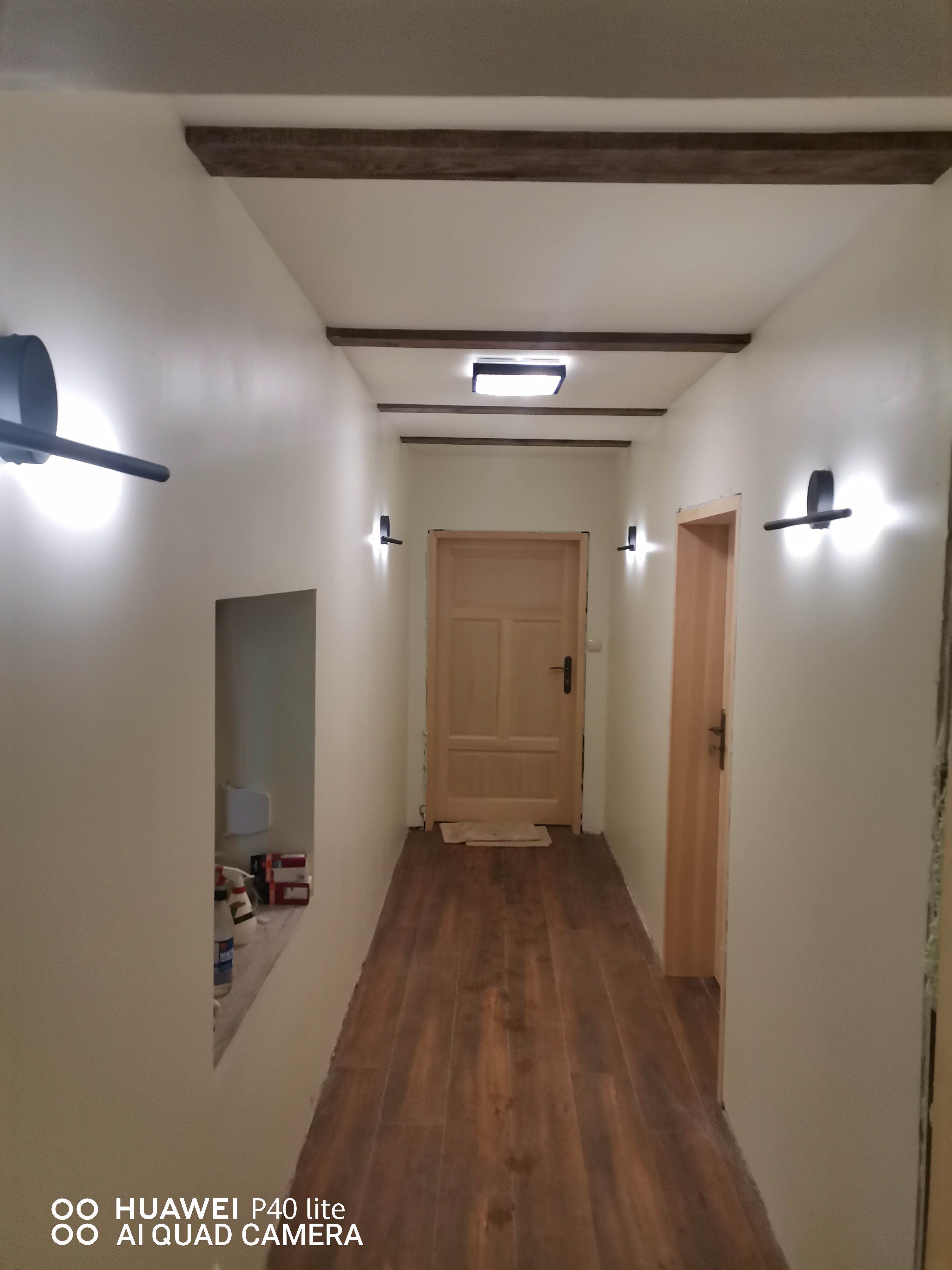 Korytarz po remoncie z drewnianymi belkami na suficie, brązowymi panelami podłogowymi, kremowymi ścianami i nowoczesnymi, czarnymi kinkietami.