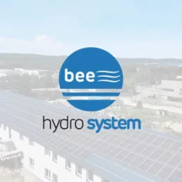 Logo firmy 'bee hydro system' na tle budynku z panelami słonecznymi na dachu, widok z lotu ptaka, lekko zamglone tło z zabudowaniami przemysłowymi.