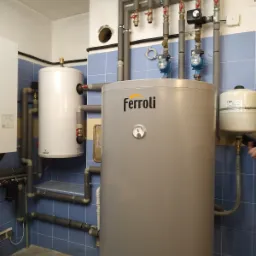 Szara, duża centrala grzewcza Ferroli z podłączonymi rurami i pompami obiegowymi w kotłowni wyłożonej niebieskimi płytkami.