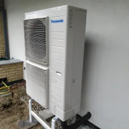 Zewnętrzna jednostka pompy ciepła Panasonic zamontowana na betonowych podstawach z białym stelażem, z czarną rurą odprowadzającą skropliny, na tle szarej ściany budynku i ziemi wysypanej kamieniami.