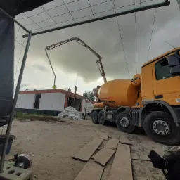 Żółta betoniarka z wysięgnikiem pompuje beton na budowę domu z białego pustaka, widoczne narzędzia i materiały budowlane, pochmurne niebo w tle.