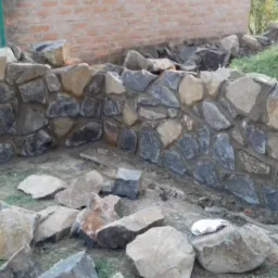 Fragment muru z kamienia polnego, z widocznym etapem budowy, surowe otoczenie placu budowy z porozrzucanymi kamieniami i fragmentami cegieł. Część muru połączona z ceglaną ścianą.