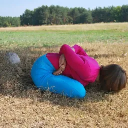 Kobieta w różowej koszulce i niebieskich spodniach wykonuje zaawansowaną pozycję zwiniętej kuli na trawie przy drzewie, z widocznymi stopami i splecionymi dłońmi za plecami.
