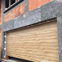PRESTIGE HOME GROUP Sp. z o.o. - Jasna, segmentowa brama garażowa z imitacją drewna, zamontowana w surowym otworze budynku w trakcie budowy. Widoczne bloczki i okno.
