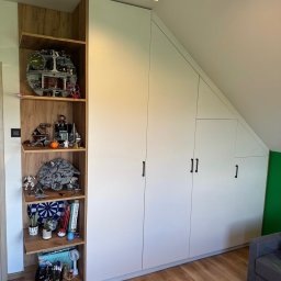 PRESTIGE HOME GROUP Sp. z o.o. - Biała szafa wnękowa z otwartymi półkami z drewnianym dekorem, wypełnionymi kolekcją figurek i przedmiotów. Ukośny sufit. Parkiet na podłodze.