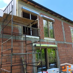 PRESTIGE HOME GROUP Sp. z o.o. - Dom w trakcie budowy z oknami i roletami zewnętrznymi, widoczne rusztowania i materiały budowlane. Rolety marki Krishome.