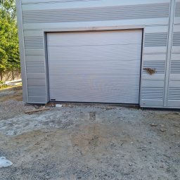 PRESTIGE HOME GROUP Sp. z o.o. - Zamknięta, segmentowa, jasnoszara brama garażowa, z poziomymi przetłoczeniami, w budynku o metalowej elewacji. Widoczne ślady prac budowlanych.