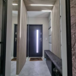 PRESTIGE HOME GROUP Sp. z o.o. - Nowoczesny przedpokój z lustrem, drewnianymi lamelami z podświetleniem LED na suficie i ścianach, szarą tapicerowaną ławką i ciemnymi drzwiami wejściowymi.