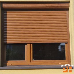 PRESTIGE HOME GROUP Sp. z o.o. - Drewniana roleta zewnętrzna, częściowo opuszczona, na oknie z brązową ramą. Jasna, teksturowana elewacja budynku w tle. Widoczne logo firmy w prawym dolnym rogu.