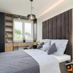 Nowoczesna sypialnia z zabudową meblową: czarna szafa, regał z jasnego drewna, biurko pod oknem i tapicerowany panel za łóżkiem. Minimalistyczny design, oświetlenie punktowe.