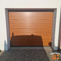 Segmentowa brama garażowa w odcieniu drewna, z widoczną fakturą, zamontowana w białej elewacji budynku. Obok rynna w kolorze miedzi.