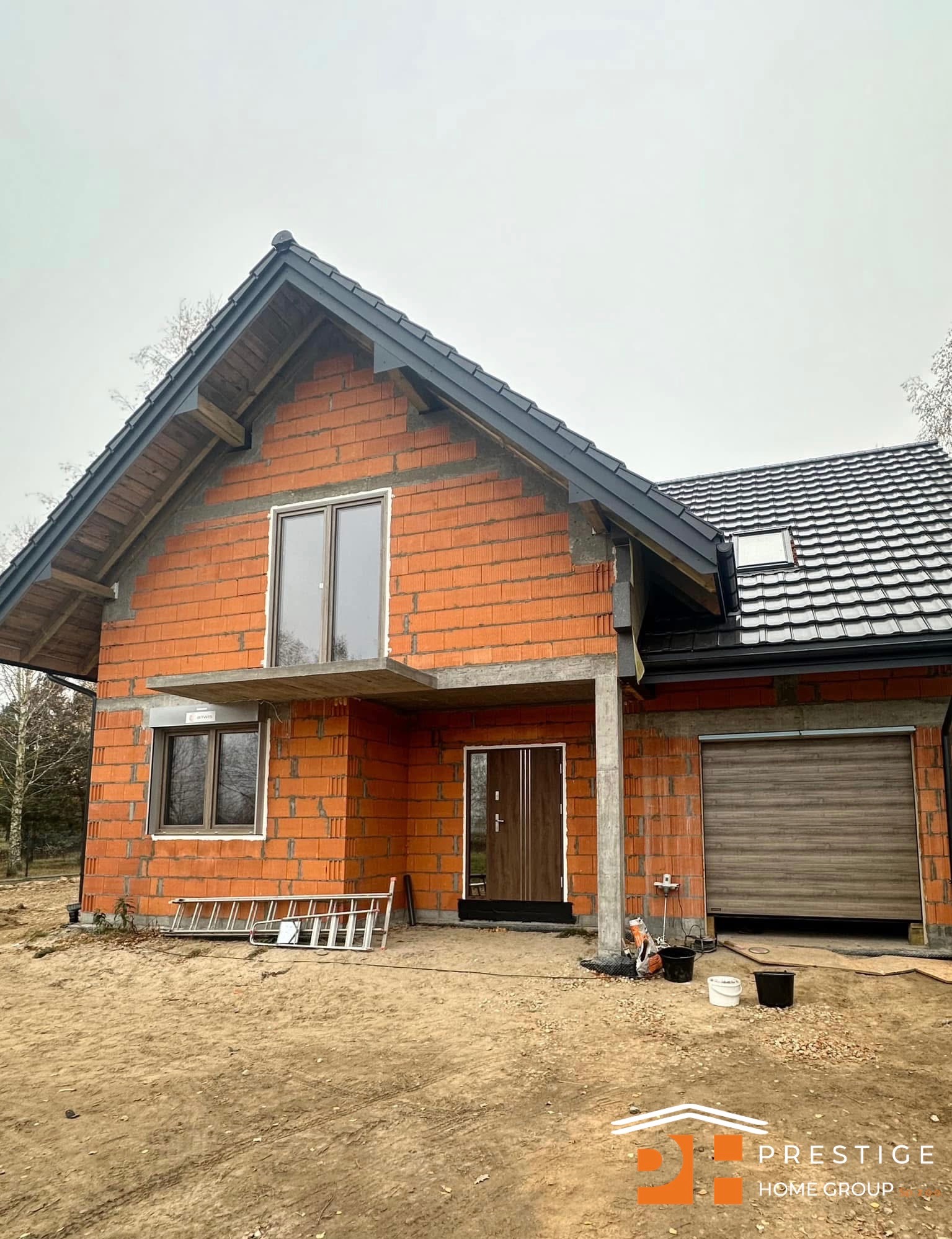 Dom w stanie surowym otwartym z cegły, z oknami PCV i bramą garażową, drabina oparta o ścianę, widoczne detale konstrukcyjne dachu i logo firmy na dole.