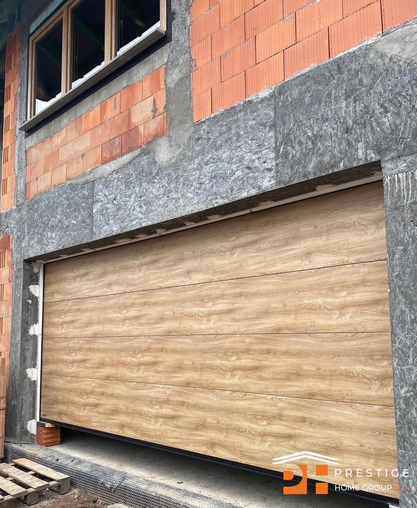 Jasna, segmentowa brama garażowa z imitacją drewna, zamontowana w surowym otworze budynku w trakcie budowy. Widoczne bloczki i okno.