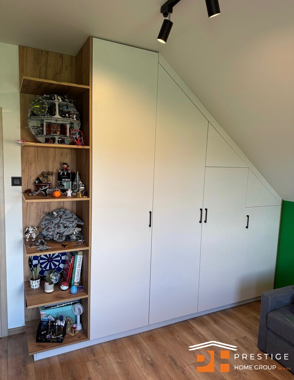 Biała szafa wnękowa z otwartymi półkami z drewnianym dekorem, wypełnionymi kolekcją figurek i przedmiotów. Ukośny sufit. Parkiet na podłodze.