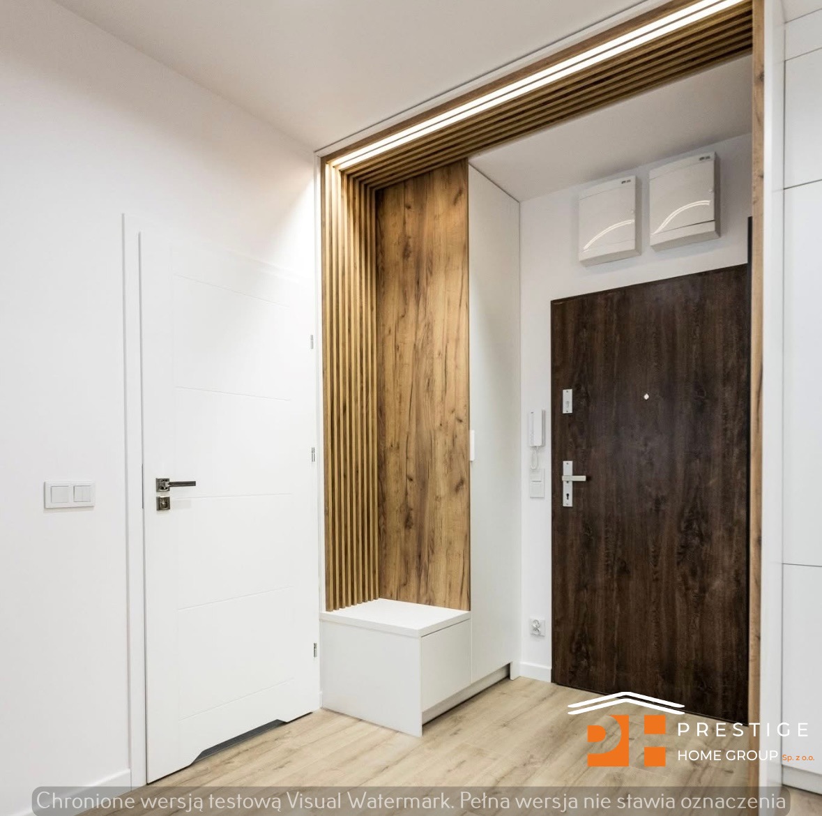 Nowoczesny przedpokój z drewnianą zabudową, oświetleniem LED i białymi drzwiami. Widoczna szafka z siedziskiem i drzwi wejściowe w kolorze ciemnego drewna.
