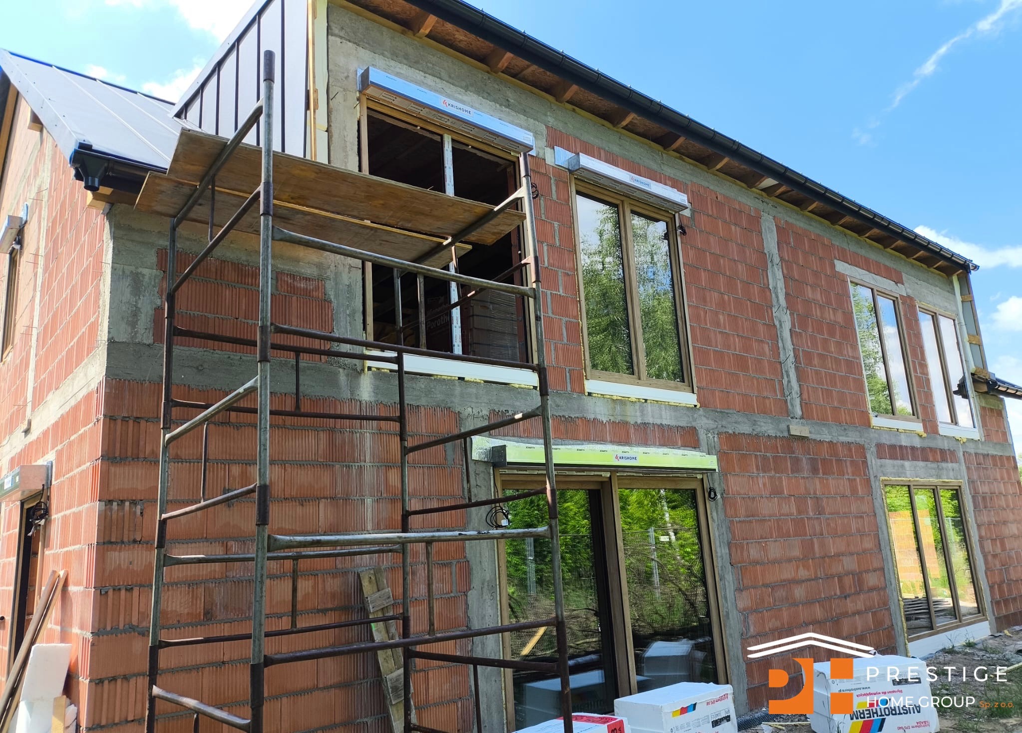 Dom w trakcie budowy z oknami i roletami zewnętrznymi, widoczne rusztowania i materiały budowlane. Rolety marki Krishome.