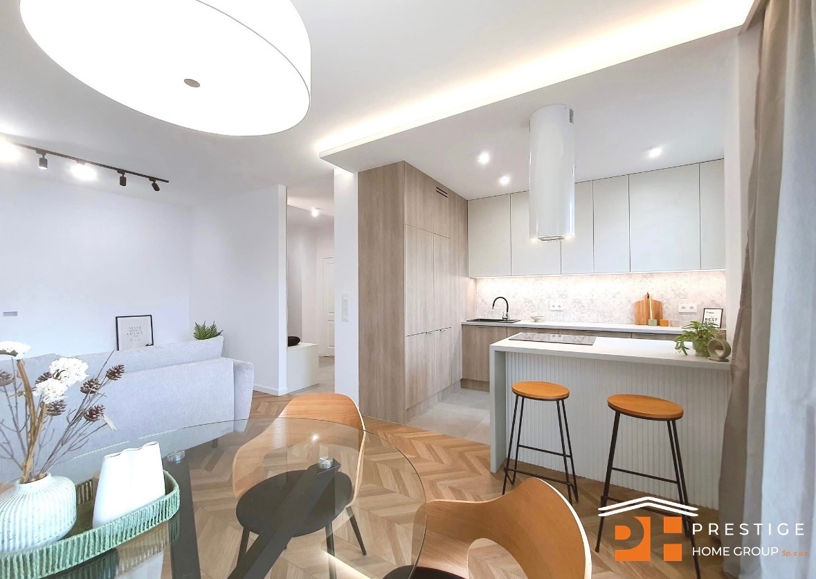 Jasna kuchnia z drewnianymi akcentami, wyspą i hokerami. Parkiet ułożony w jodełkę. W tle salon z szarą sofą i dekoracjami. Całość w nowoczesnym stylu.