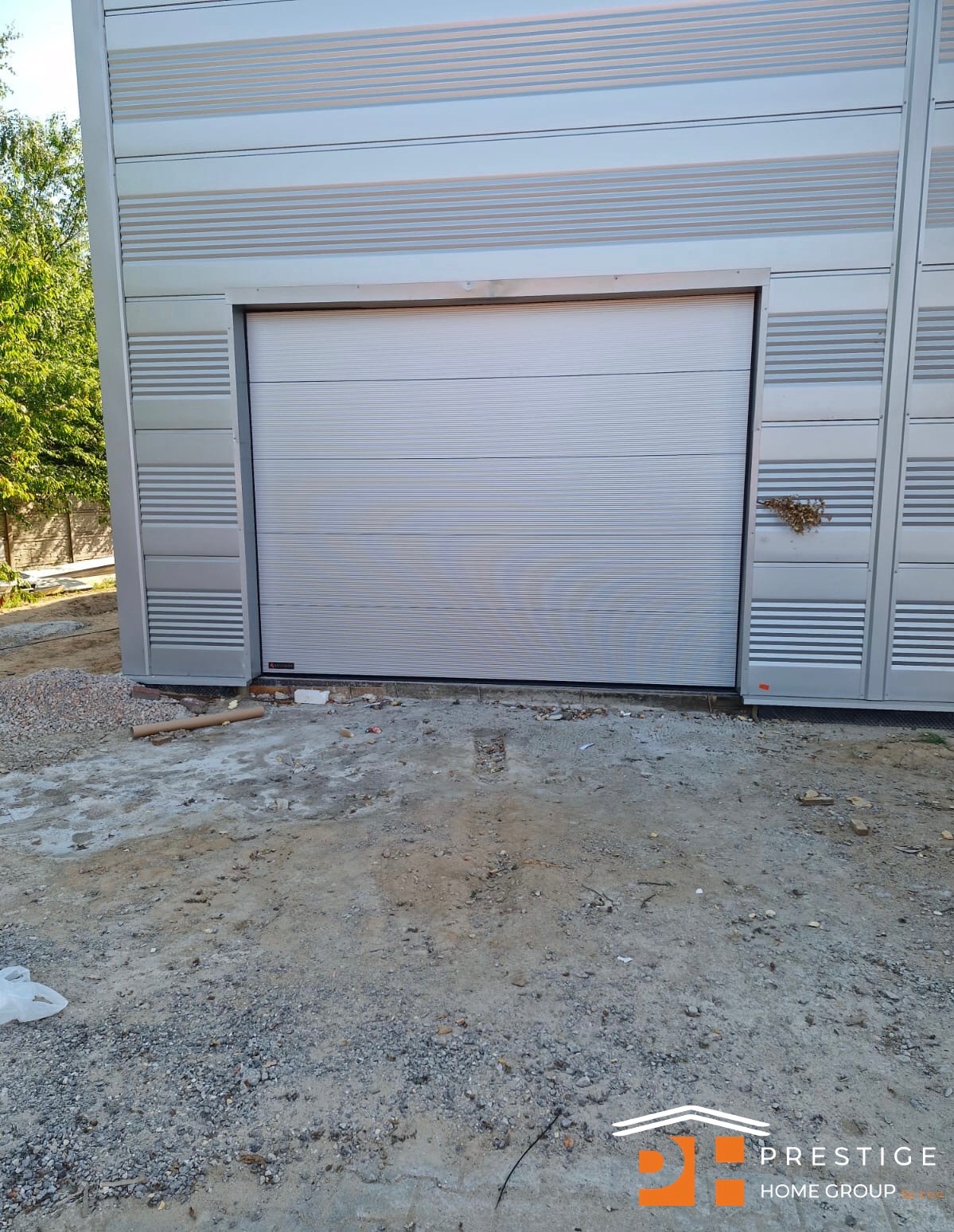 Zamknięta, segmentowa, jasnoszara brama garażowa, z poziomymi przetłoczeniami, w budynku o metalowej elewacji. Widoczne ślady prac budowlanych.