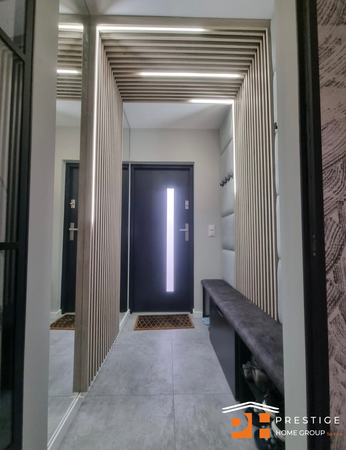 Nowoczesny przedpokój z lustrem, drewnianymi lamelami z podświetleniem LED na suficie i ścianach, szarą tapicerowaną ławką i ciemnymi drzwiami wejściowymi.