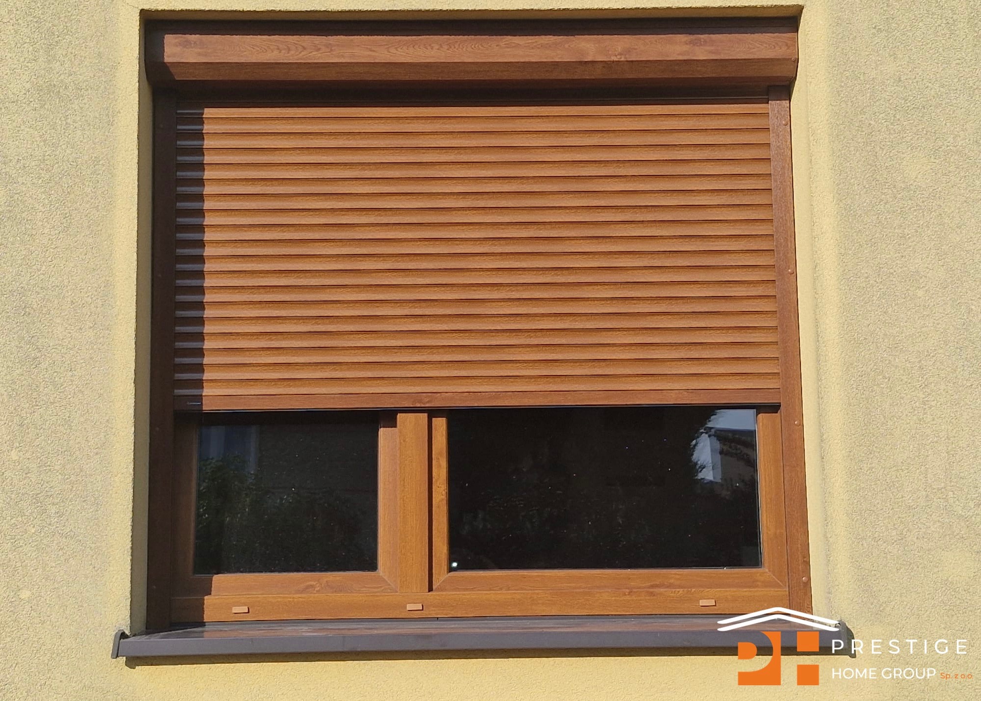 Drewniana roleta zewnętrzna, częściowo opuszczona, na oknie z brązową ramą. Jasna, teksturowana elewacja budynku w tle. Widoczne logo firmy w prawym dolnym rogu.