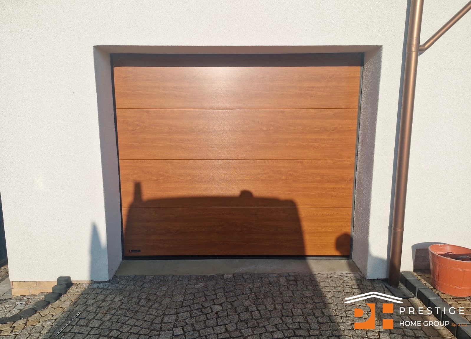 Segmentowa brama garażowa w odcieniu drewna, z widoczną fakturą, zamontowana w białej elewacji budynku. Obok rynna w kolorze miedzi.