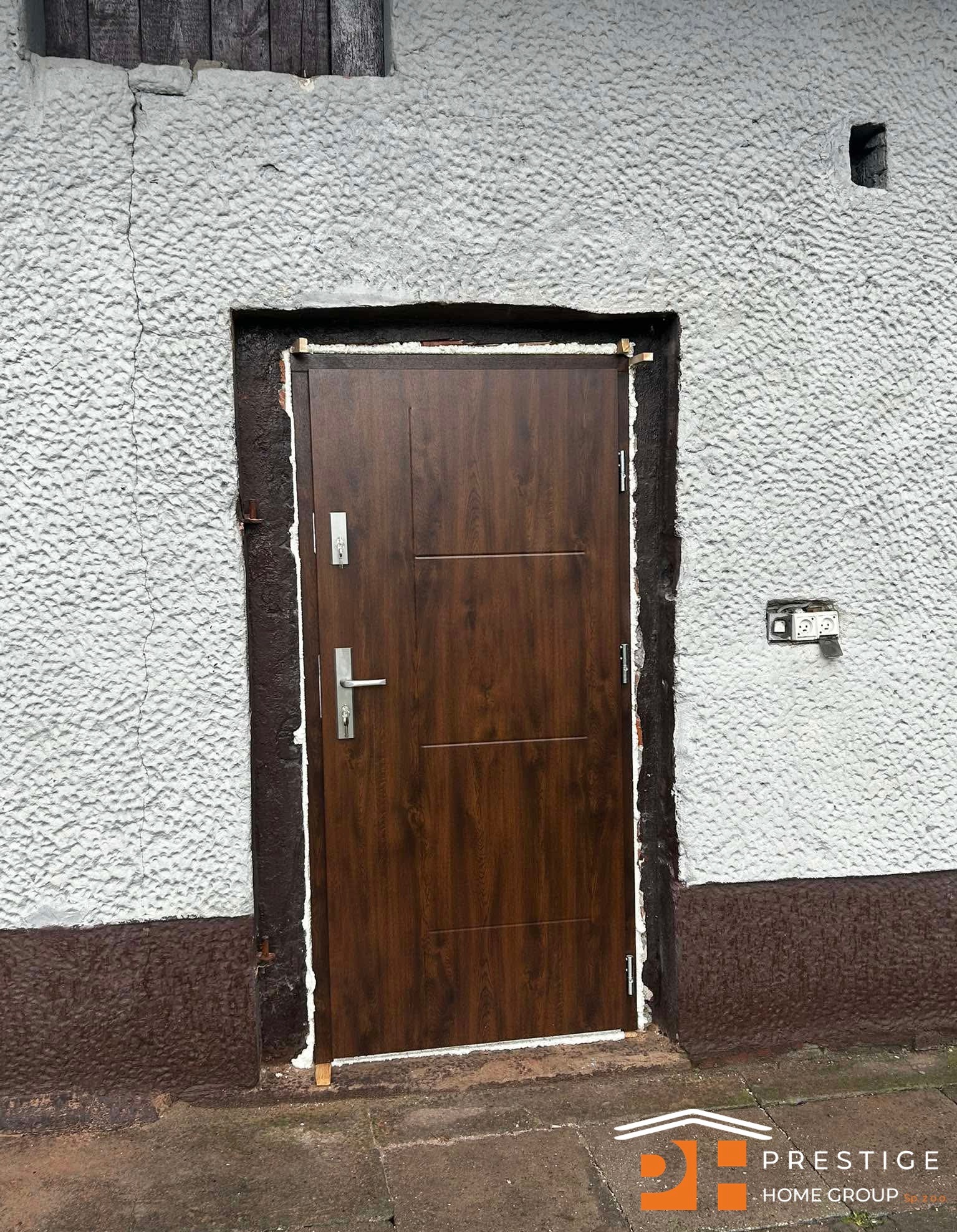 Brązowe drzwi wejściowe z widoczną ościeżnicą i pianką montażową, osadzone w szarej, chropowatej ścianie budynku. Widoczny fragment elewacji i gniazdko elektryczne.