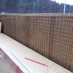 Szczegółowe ujęcie zbrojenia pionowej ściany fundamentowej w szalunku z płyty OSB, widoczne poziomice i narzędzia na podłożu, przygotowanie do betonowania.