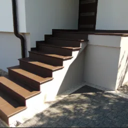 Zewnętrzne schody wejściowe wyłożone brązowymi płytkami ceramicznymi, z widocznym fragmentem elewacji budynku i kostką brukową na podjeździe, oświetlone popołudniowym słońcem.