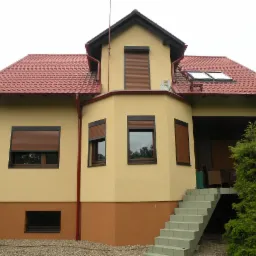 Dwukondygnacyjny dom z żółtą elewacją, brązowymi roletami i czerwoną dachówką, widoczne schody prowadzące na taras.