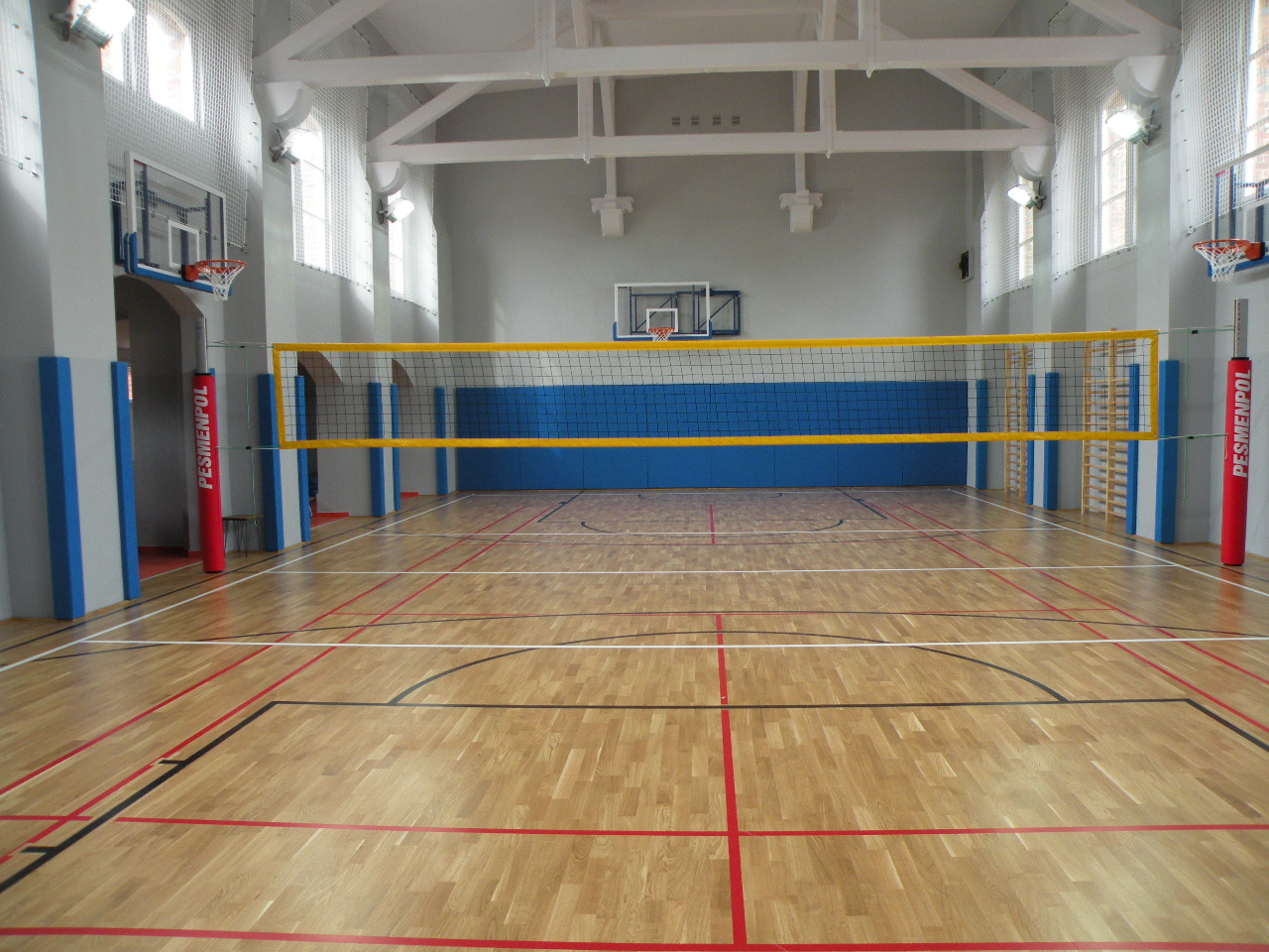 Jasna sala gimnastyczna z parkietem w odcieniach brązu, siatką do siatkówki z żółtymi taśmami i koszami do koszykówki po bokach, oświetlona dużymi oknami.