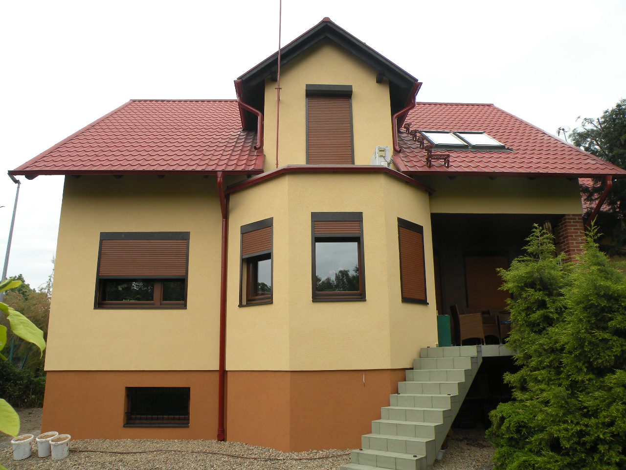 Dwukondygnacyjny dom z żółtą elewacją, brązowymi roletami i czerwoną dachówką, widoczne schody prowadzące na taras.