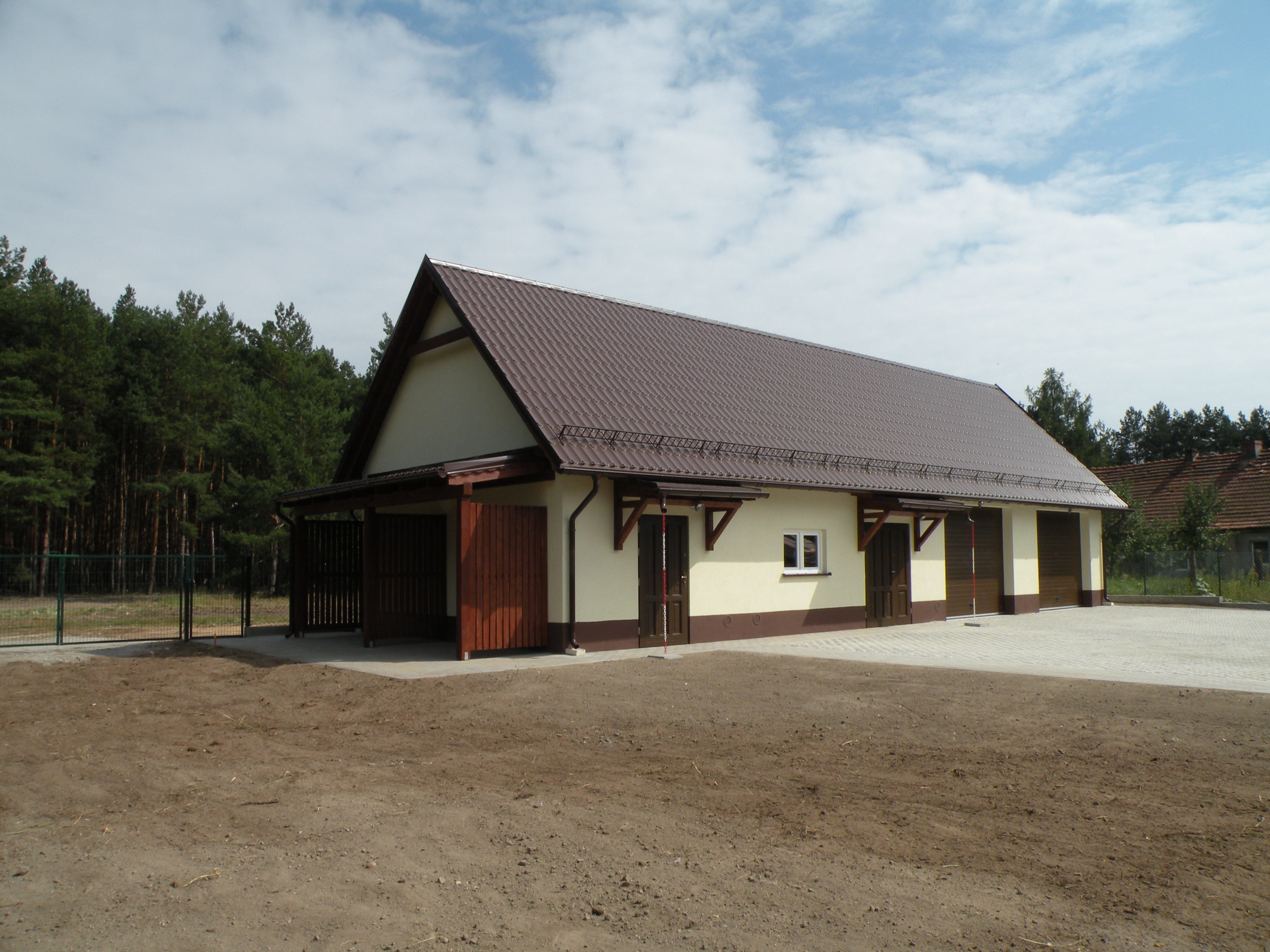 Nowo wybudowany, parterowy budynek gospodarczy z brązowym dachem z blachodachówki, beżową elewacją z brązowymi akcentami, drewnianymi wrotami garażowymi i ogrodzeniem z siatki na tle lasu.