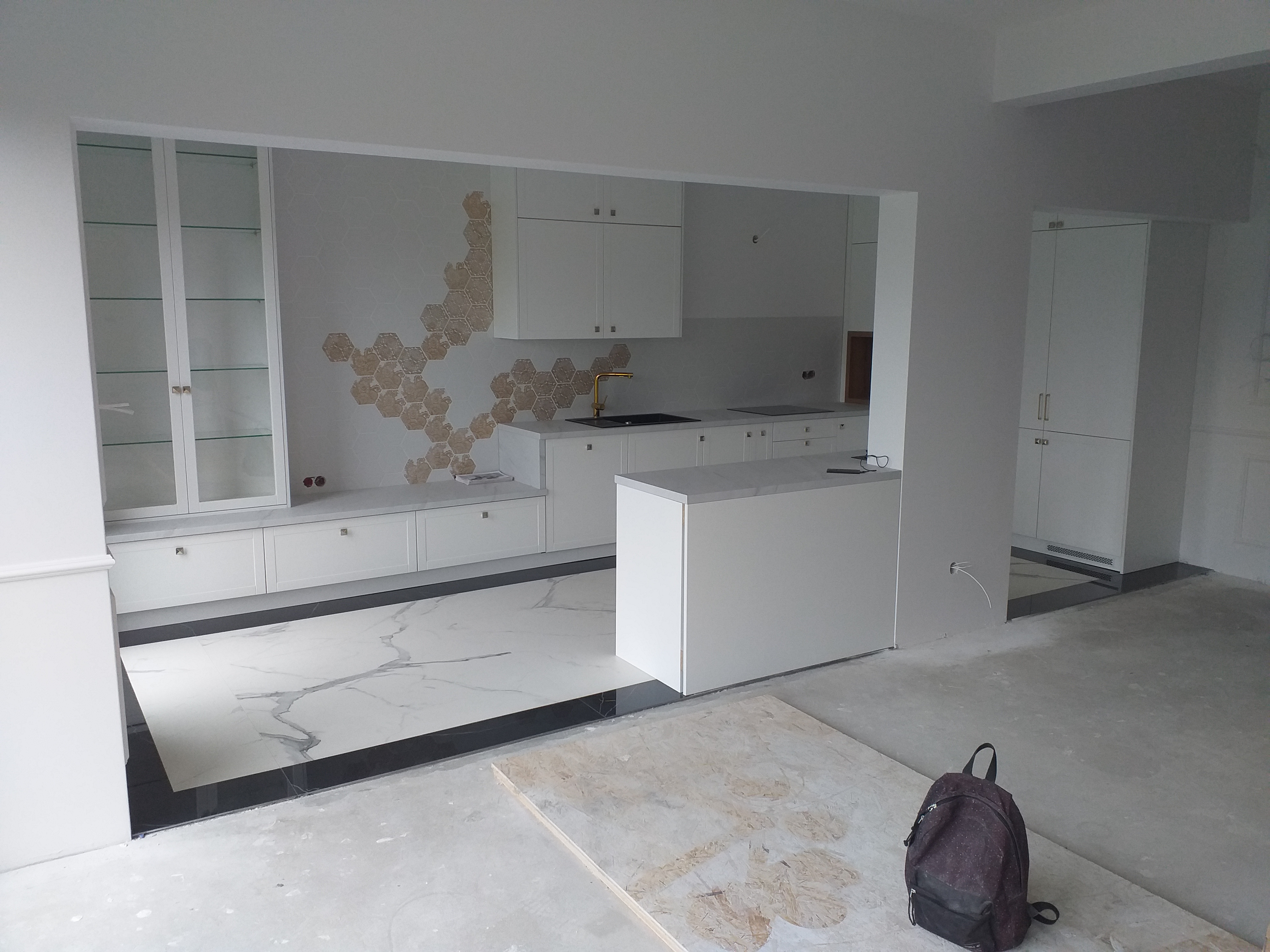 Wnętrze kuchni w trakcie prac wykończeniowych: białe szafki, marmurowa podłoga z czarną obwódką, dekoracyjna mozaika heksagonalna na ścianie, plecak leżący na podłodze.