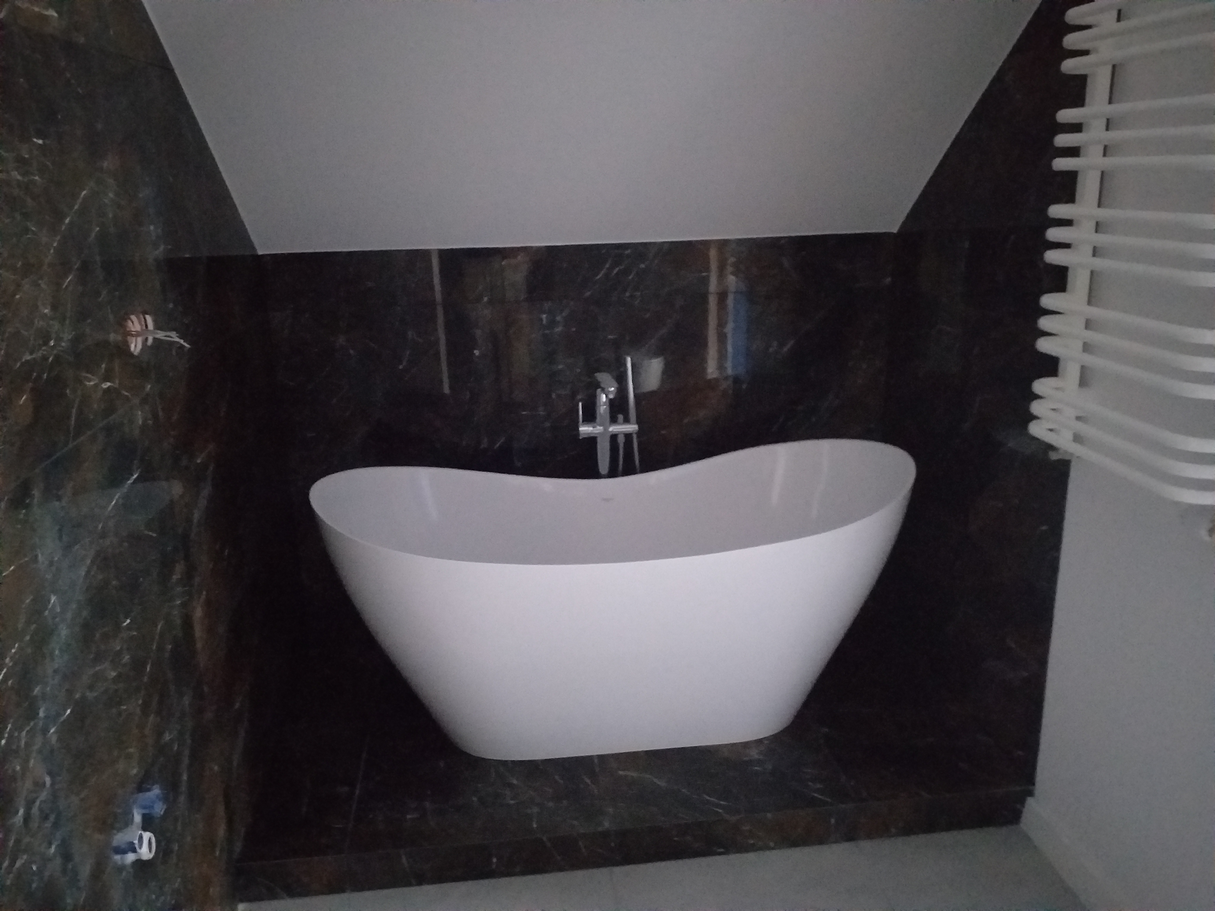 Wolnostojąca, biała wanna o opływowym kształcie z chromowaną armaturą na tle ściany wyłożonej ciemnym marmurem z wyraźnym użyleniem, obok grzejnik drabinkowy.