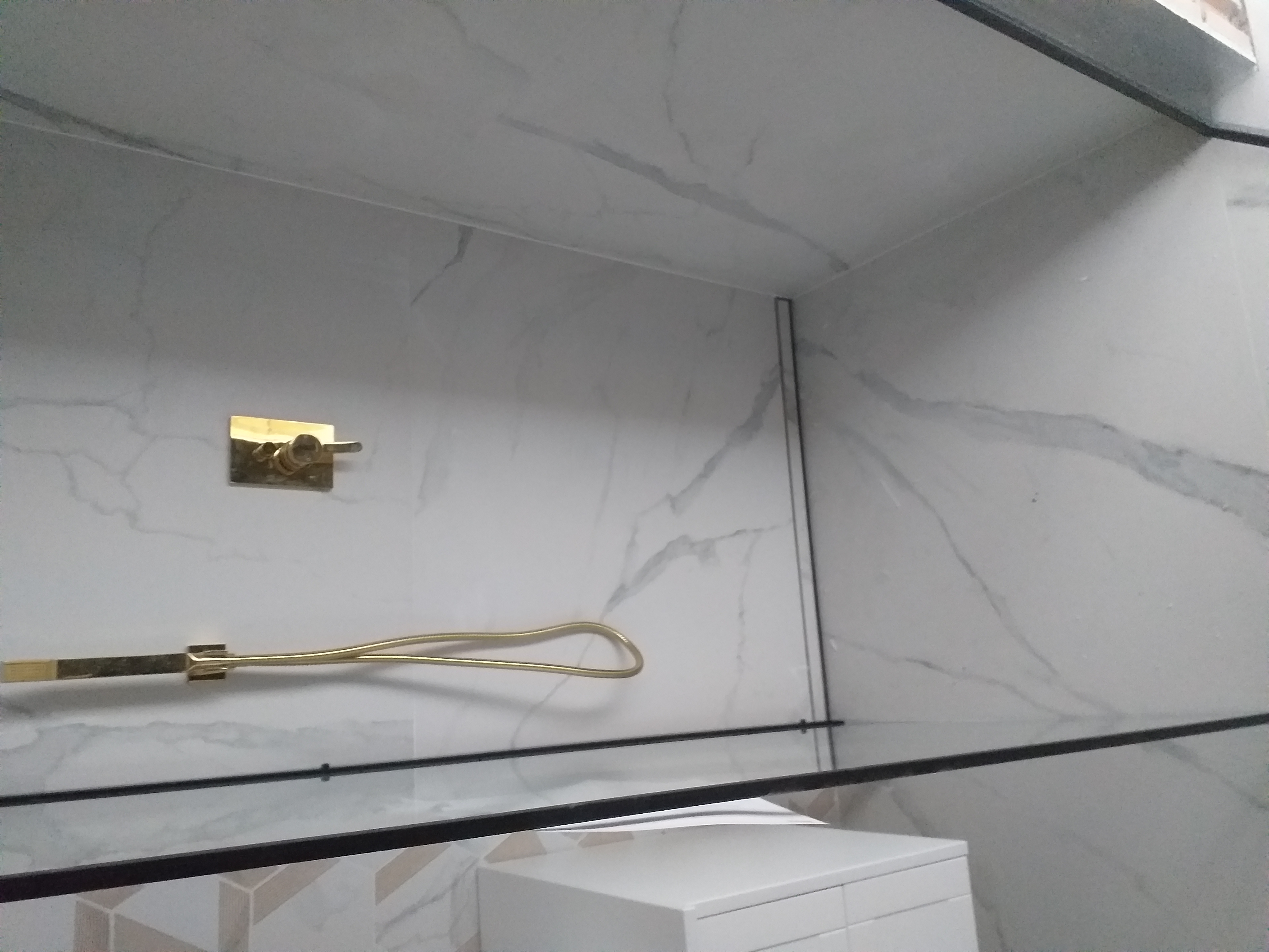 Biała łazienka z marmurowymi płytkami, złotym uchwytem na ręczniki i baterią, oraz czarną szklaną półką nad białą szafką pod umywalką.