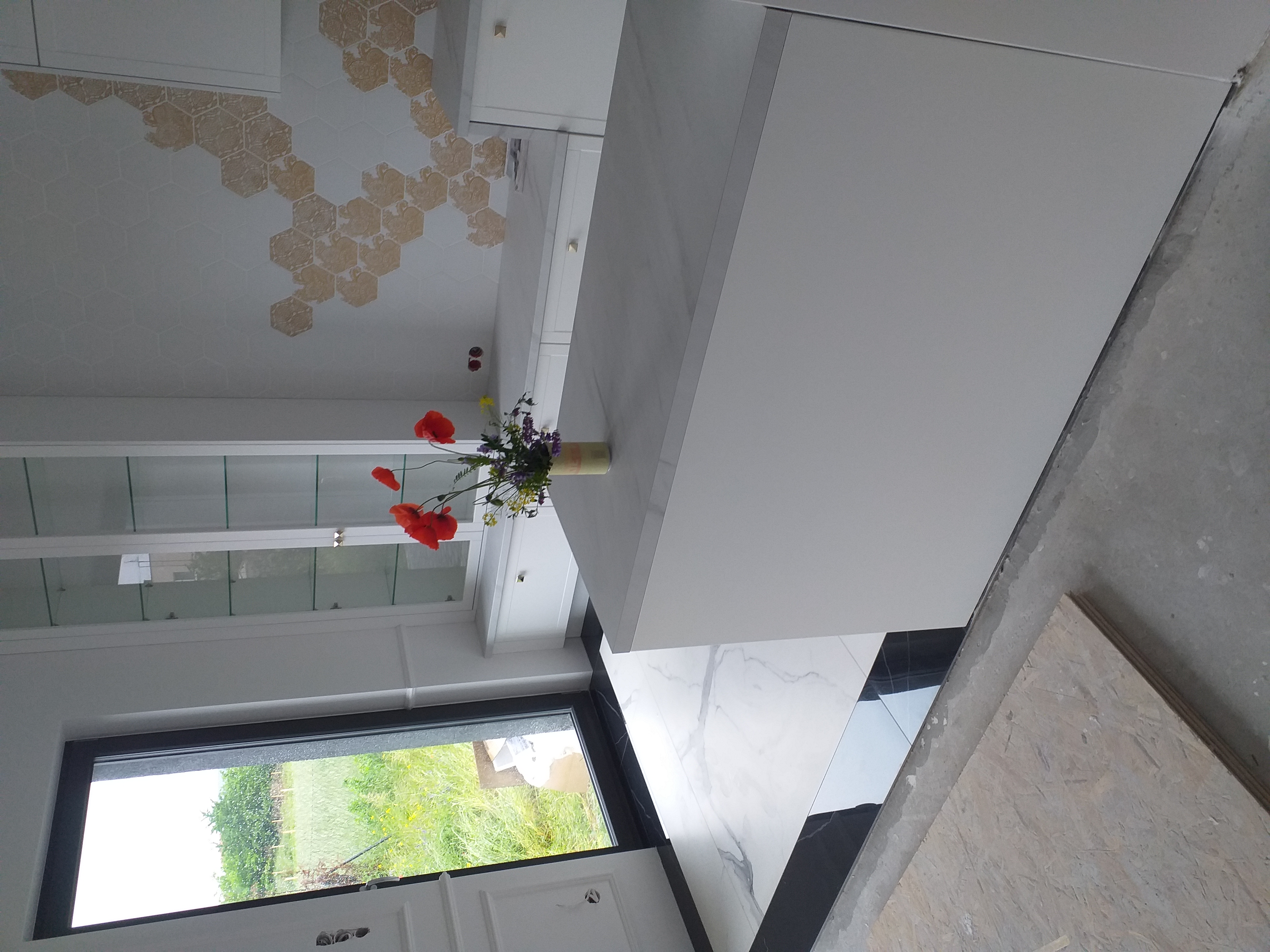 Nowoczesna kuchnia w trakcie wykończenia: białe szafki, marmurowe płytki podłogowe, dekoracyjna mozaika heksagonalna na suficie, bukiet czerwonych maków na wyspie kuchennej, widok na zieleń za oknem.