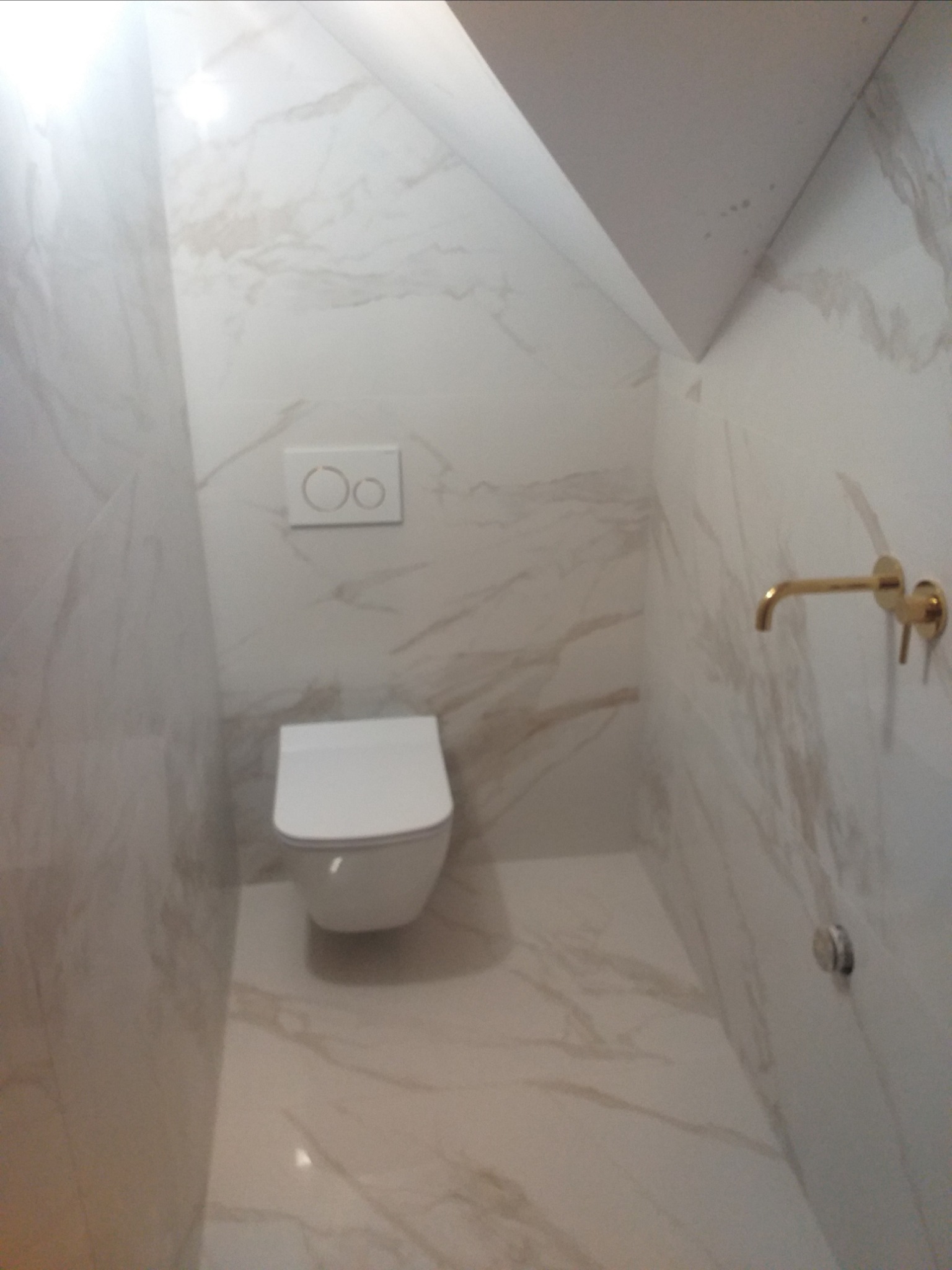 Nowoczesna toaleta w małej łazience wykończonej płytkami imitującymi marmur, z podtynkową spłuczką i złotą baterią umywalkową.