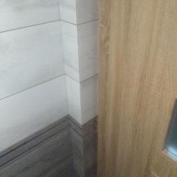 J&J - Wykończony narożnik łazienki z jasnymi płytkami ściennymi i ciemniejszymi płytkami podłogowymi imitującymi drewno, przylegający do drewnianej futryny drzwiowej.