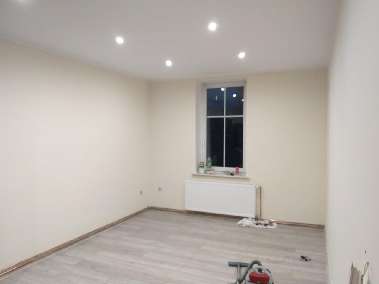 Wnętrze pokoju po malowaniu ścian na kremowo, z widoczną listwą przypodłogową, grzejnikiem pod oknem i jasnymi panelami podłogowymi. Na suficie zamontowane punktowe oświetlenie.